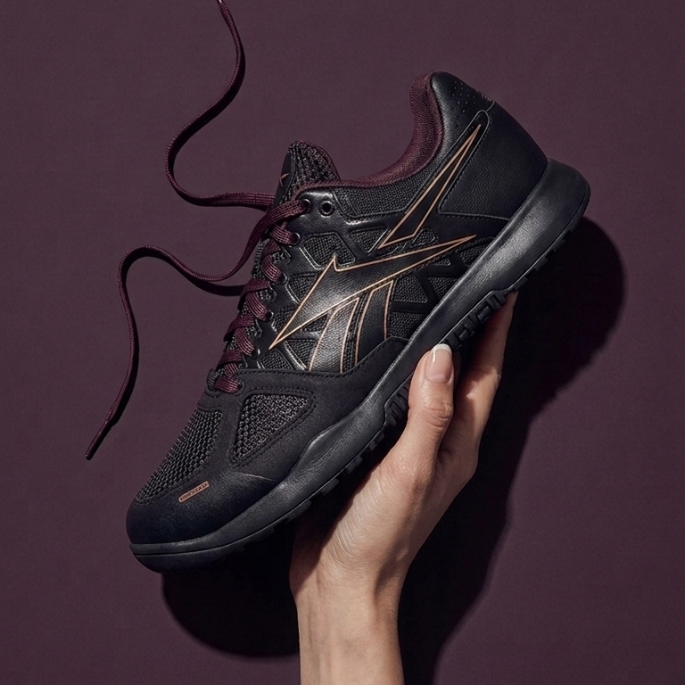 A black Reebok Nano 2.0 with gold side stripes and burgundy laces, held in a person's hand against a dark purple background.